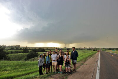 Students standing on the side of the road posing for the camera in front of a cloud formation and tornado