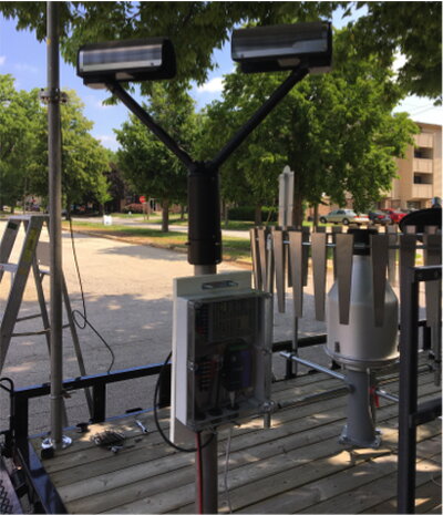 Outdoor monitoring station with two mounted cameras and weather instruments on a platform near a street.