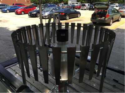 Circular rain gauge with metal wind shield mounted on a platform in a parking lot.