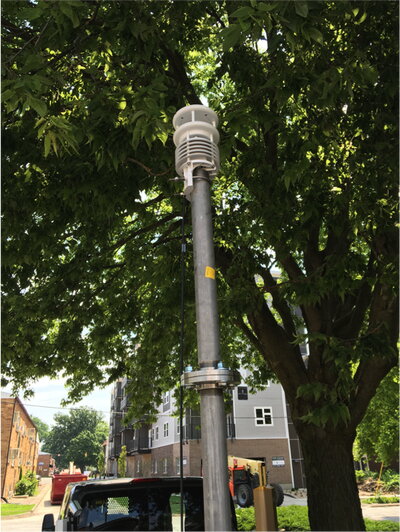 Air temperature and humidity sensor mounted on a pole beneath a tree canopy near residential buildings.